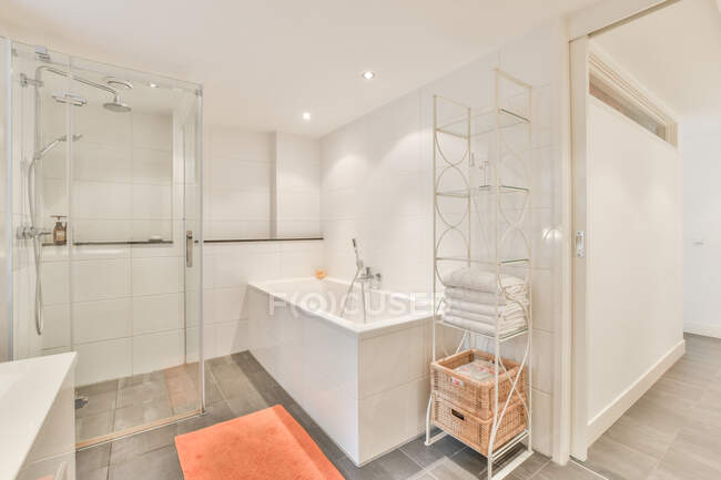 Moderna vasca da bagno e cabina doccia vicino a scaffali con asciugamani e cestini in bagno contemporaneo con pareti bianche — Foto stock