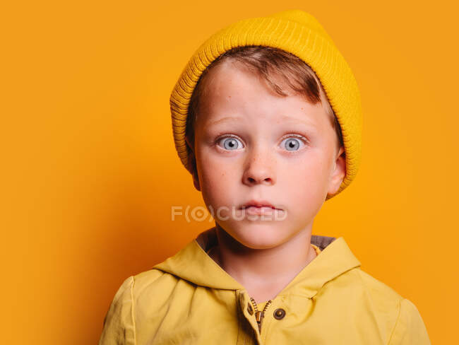 Niño asombrado con ojos azules en chaqueta de otoño de color amarillo brillante y sombrero de gorro mirando a la cámara contra el fondo amarillo en el estudio - foto de stock