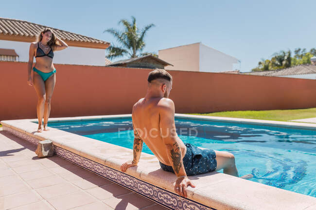 Любящие пары в купальниках, отдыхающие рядом с бассейном рядом со зданием в солнечный летний день в тропическом курорте во время отпуска — стоковое фото