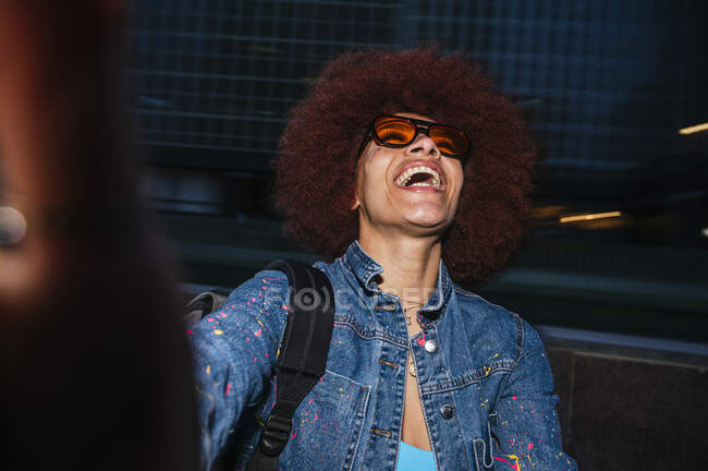 Joyeuse femme avec une coiffure afro portant une tenue denim tendance et des lunettes de soleil prenant autoportrait dans la rue sombre en soirée — Photo de stock
