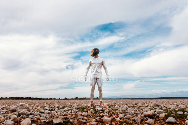 Masculino em máscara de macaco e roupa de látex de prata em pé no campo pedregoso e olhando para longe contra o céu azul nublado — Fotografia de Stock
