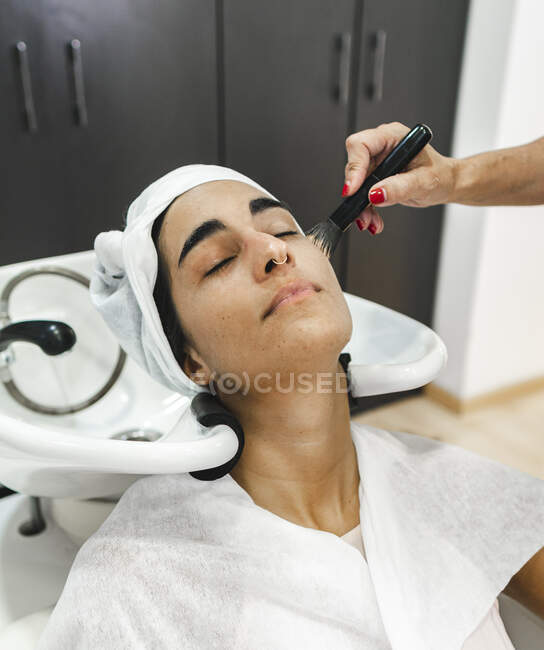 From above of crop unrecognizable female master using brush for making makeup woman with closed eyes after washing hair — Stock Photo