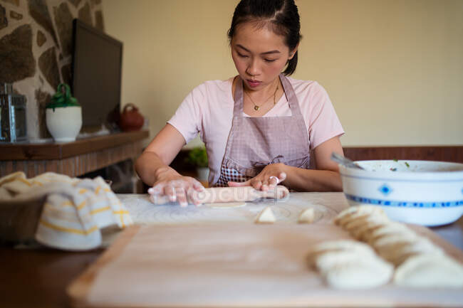 Снизу женщина в фартуке катит тесто на столе, пока готовит пельмени с мясом на кухне — стоковое фото