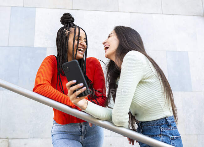 From below of cheerful diverse females in stylish clothes standing on stairs near railing and laughing while browsing smartphone on street in daytime — Stock Photo