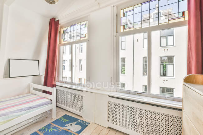 Interior do quarto leve mobilado com cama confortável e armário de madeira perto de janelas com cortinas à luz do dia — Fotografia de Stock