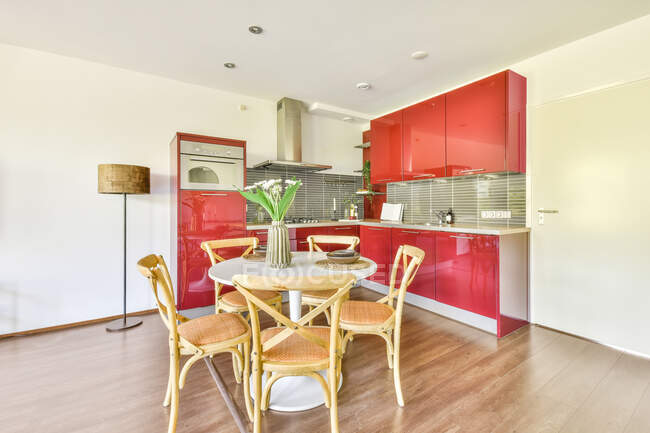 Intérieur moderne de cuisine avec armoires rouges et table à manger blanche décorée de fleurs dans un vase dans un appartement contemporain — Photo de stock