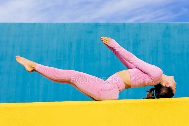 Full body of slim barefoot woman balancing in variation of Matsyasana pose with raised legs and hands on yellow and blue wall — Stock Photo