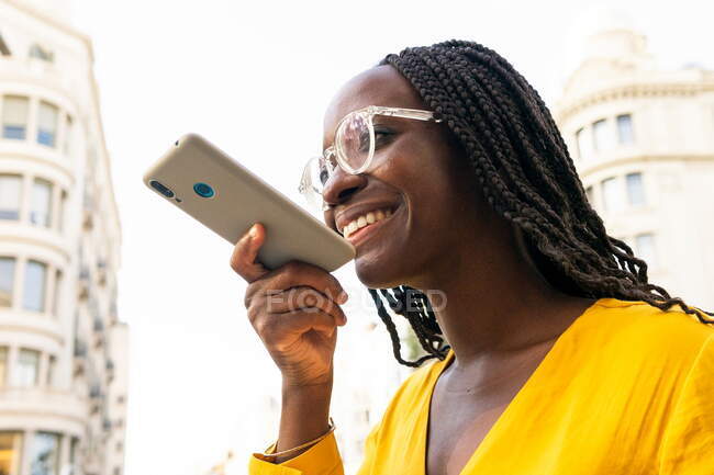 Positive African American female with black hair in sunglasses recording voice message on modern smartphone while strolling through the city streets — Stock Photo