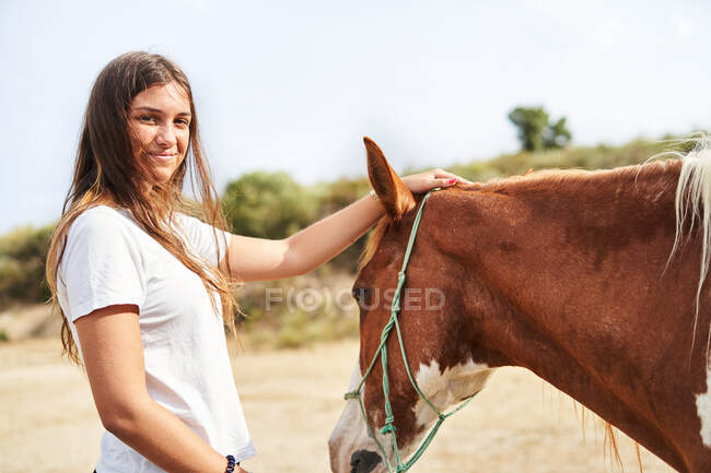 Mujer feliz acariciando caballo con brida en la mano mientras está de pie en el suelo arenoso cerca de la barrera y las plantas a la luz del día en la granja mirando a la cámara - foto de stock