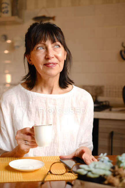 Mujer positiva de mediana edad sonriendo brillantemente mientras disfruta de bebidas calientes en la cocina por la mañana y mirando hacia otro lado - foto de stock