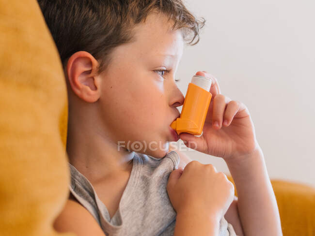 Вид сбоку маленького мальчика с астмой, который использует ингалятор, сидя дома на диване — стоковое фото