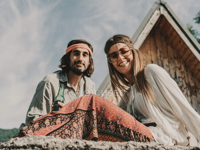 From below positive hippie couple in boho styled clothes sitting near wooden structure during trip in nature — Stock Photo
