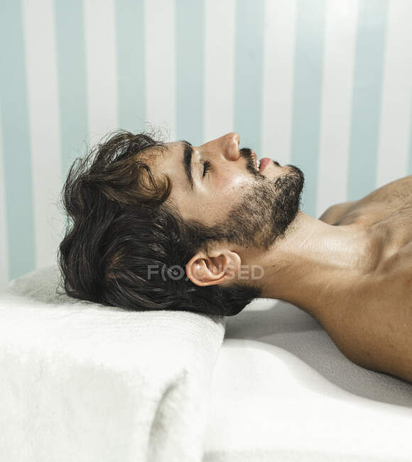 Vista lateral del hombre barbudo sin camisa acostado en la cama de masaje con los ojos cerrados - foto de stock