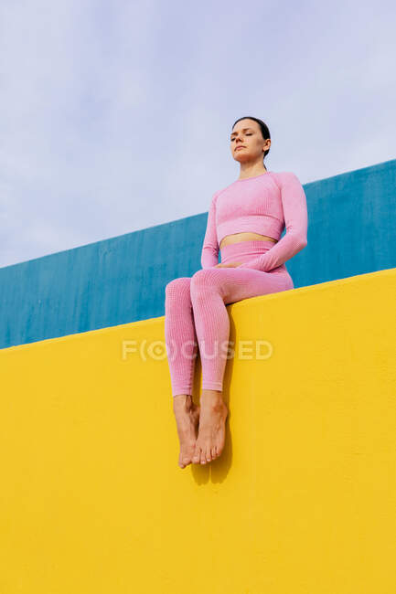 Low angle full body of confident fit female in pink sportswear sitting with closed eyes on bright yellow surface — Stock Photo