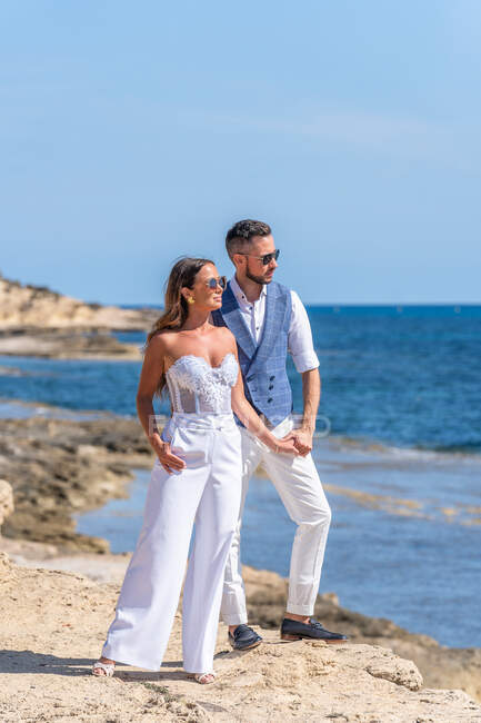 Full body of young couple in elegant outfit and sunglasses standing near sea on hill on rocky coast while holding hands and looking away under blue sky in sunny day — Stock Photo