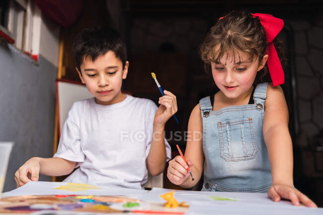Niños positivos con pinceles pintura con acuarelas de colores sobre papel en la mesa - foto de stock