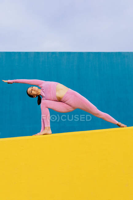 Slim female in pink sportswear practicing yoga in Utthita Parshvakonasana on bright blue and yellow background — Stock Photo