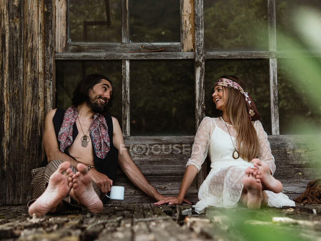 Hippie pareja sentada en el suelo del porche mirándose mientras toma café - foto de stock
