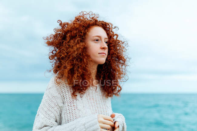 Thoughtful female with curly long ginger hair wearing knitted sweater standing looking away against blue sea — Stock Photo