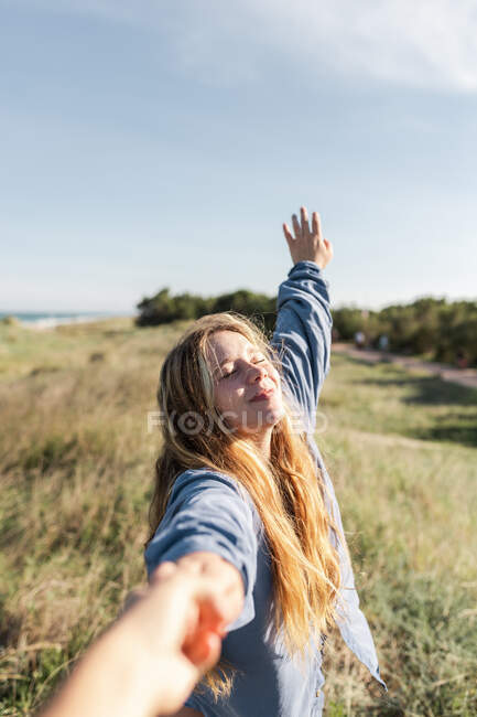 Щаслива жінка в повсякденному одязі тримає руку врожаю невпізнаваного партнера, стоячи з закритими очима на трав'янистому полі — стокове фото
