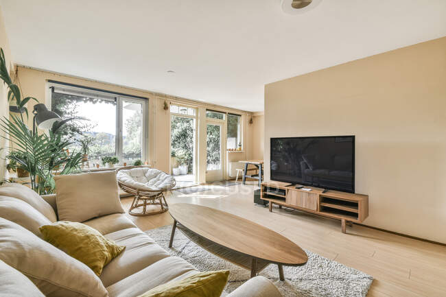 Interior de la luz sala de estar con ventanas francesas sillón de ratán sofá beige con cojines flores interiores y madera oval mesa TV y alfombra en el suelo - foto de stock