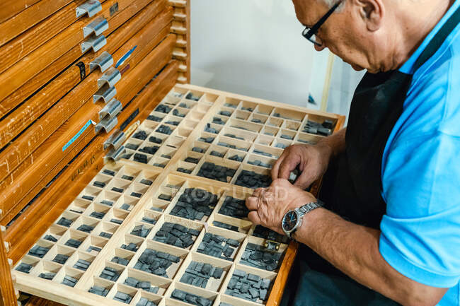 Сверху на урожае сконцентрирован старший ремесленник мужского пола в фартуке и очках, выбирающий печатные буквы из деревянной коробки во время работы в традиционном ателье — стоковое фото