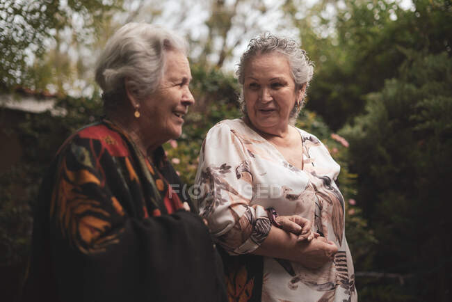 Alte Damen in lässiger Kleidung unterhalten sich bei einem gemeinsamen Spaziergang im Sommergarten in der Nähe grüner Rosenbüsche an bewölkten Tagen — Stockfoto
