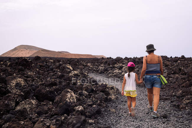 Back view of anonymous mom with girl holding hands while strolling on pathway between lava against Caldereta and Caldera Blanca Volcanoes in Spain — Stock Photo