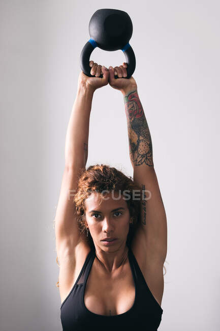 Concentre-se forte mulher fisiculturista com tatuagens levantando pesado kettlebell formação branca no ginásio contra a parede leve — Fotografia de Stock