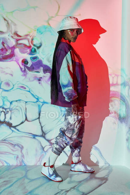 Side view of self assured young Dominican female teenager in trendy outfit and hat standing near white wall with creative abstract projections and looking at camera — Stock Photo