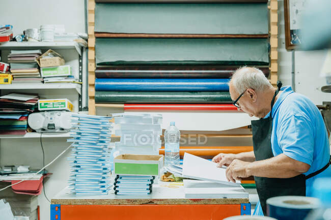 Vista lateral do artesão masculino sénior concentrado com cabelos brancos em avental e óculos em pé à mesa com pilha de papéis e caderno na oficina de impressão — Fotografia de Stock