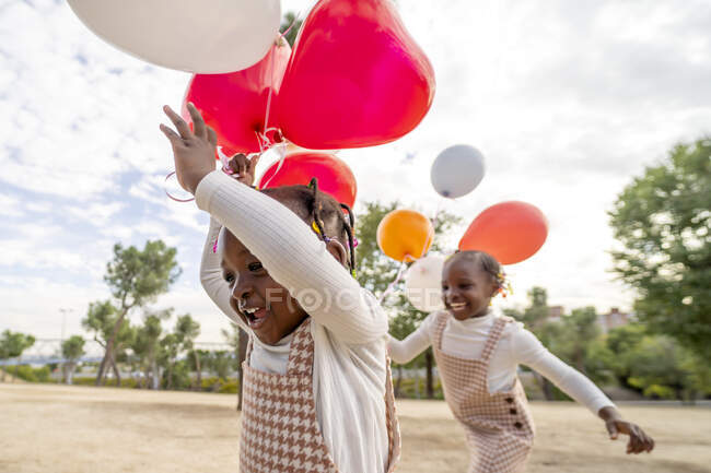 Happy African American sorelline in abiti simili in piedi con palloncini colorati in mano su erba verde in parco alla luce del giorno — Foto stock