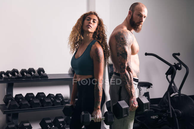 Strong sportswoman in activewear standing with dumbbells near muscular bodybuilder during workout in gym in daytime — Stock Photo