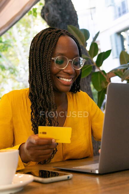 Positive African American female freelancer with credit card sitting at table with netbook while doing online purchase on terrace in cafe — Stock Photo