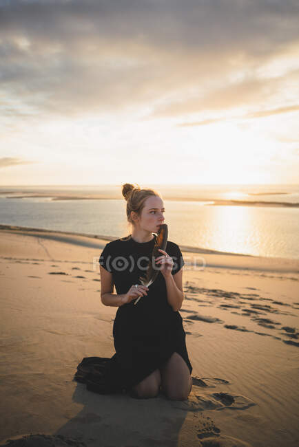 Seriöses Weibchen mit schwarzer Feder in einem Kleid, das auf einer Sanddüne kniet, die bei Sonnenuntergang vom Meer gewaschen wird — Stockfoto