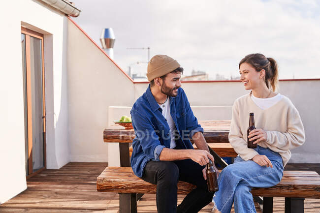 Positive junge männliche und weibliche Mitbewohner sitzen mit Bierflaschen auf Bank und unterhalten sich auf der Terrasse — Stockfoto