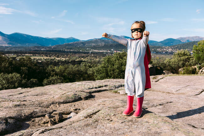 Ganzkörper eines kleinen Mädchens im Superheldenkostüm, das ausgestreckte Fäuste erhebt, um Macht zu zeigen, während es auf einem felsigen Hügel steht — Stockfoto