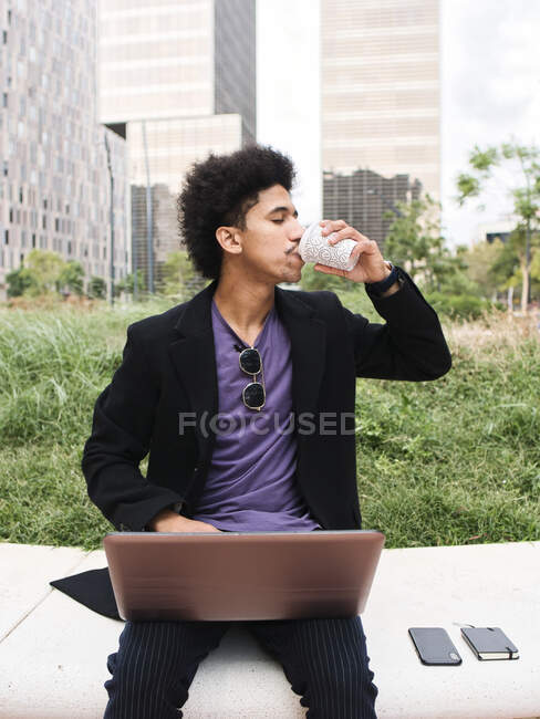 Young African American male freelancer with dark curly hair in stylish clothes drinking takeaway coffee while working remotely on laptop sitting on stone bench in city park — Stock Photo