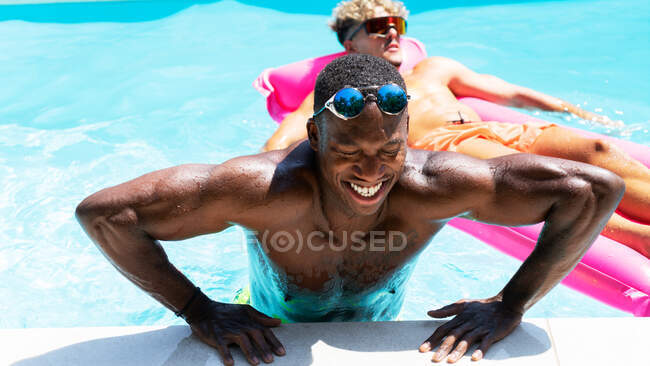 Hombre americano africano alegre con los anteojos que salen de la piscina cerca de amigo masculino en colchón inflable en día soleado del verano - foto de stock