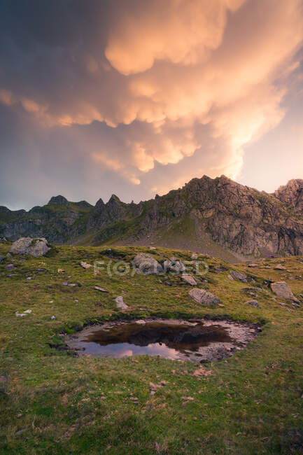 Campo erboso con pozzanghera rotonda e massi situati contro la catena montuosa rocciosa e cielo nuvoloso nella natura selvaggia della Spagna — Foto stock