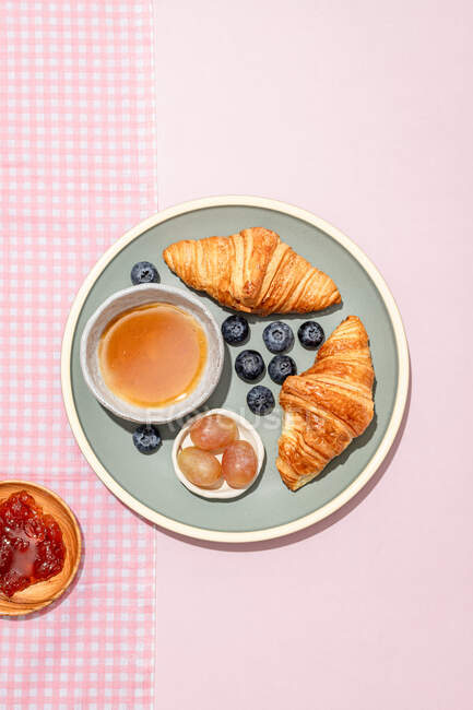 Vue de dessus de délicieux croissants servis sur une assiette en céramique avec des bleuets frais et de la confiture placée sur une table rose — Photo de stock