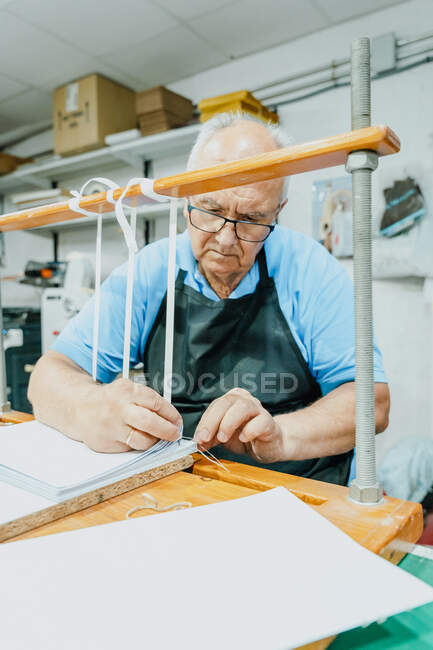 Artesanato masculino sênior atento em avental e óculos amarrando fitas em placa de madeira antes de trabalhar na máquina de impressão — Fotografia de Stock