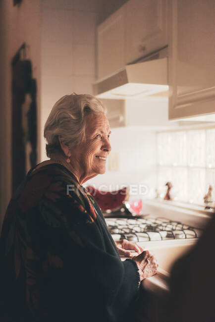 Vista laterale della felice donna anziana positiva con i capelli grigi che indossa vestiti caldi in piedi al lavandino in cucina — Foto stock