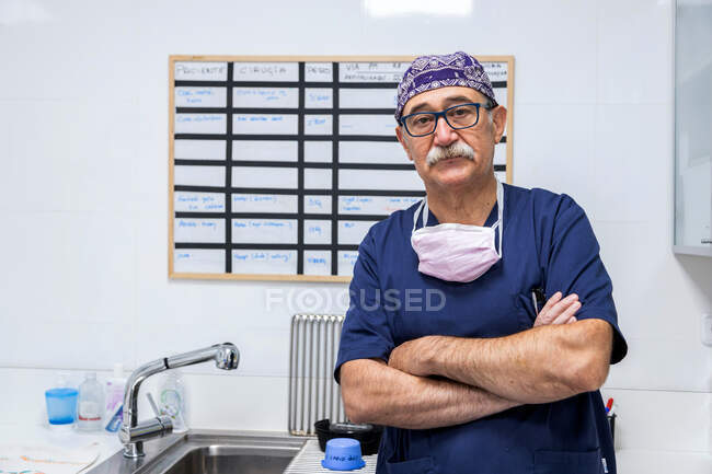 Serious old male doctor in medical uniform and hat standing with folded hands and looking at camera — Stock Photo