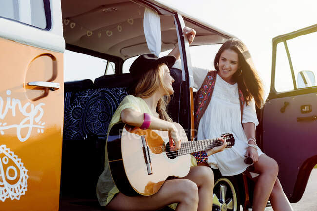 Дві красиві дівчата, одягнені в літній одяг, розважаються поза фургоном і грають на гітарі — стокове фото