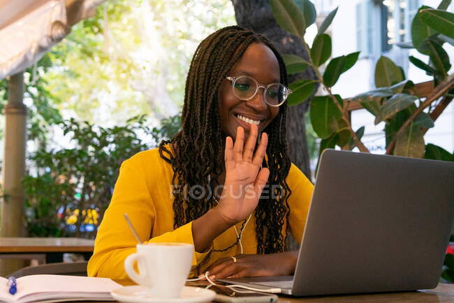 Positive African American female waving hand while having video chat on laptop at table with drink on terrace in cafeteria — Stock Photo