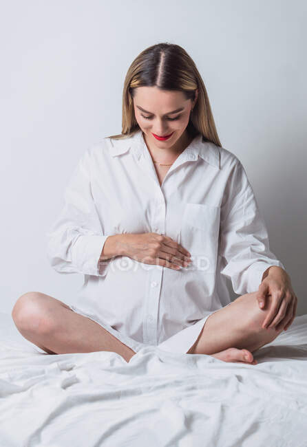 Vorderansicht einer jungen, sanft schwangeren Frau, die ihren Bauch berührt, während sie auf dem Bett sitzt und glücklich lächelt — Stockfoto
