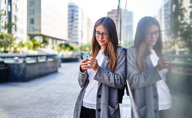 Mulher positiva em desgaste formal usando celular enquanto se inclina no prédio na rua da cidade para o trabalho — Fotografia de Stock