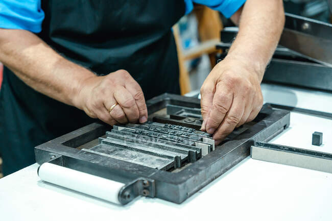 Crop unrecognizable elderly male artisan in apron and eyeglasses preparing metal frame for letterpress printing in studio — Stock Photo
