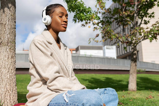 Tranquila mujer afroamericana con los ojos cerrados escuchando música en auriculares inalámbricos mientras está sentada en el césped cerca del tronco del árbol en el soleado parque - foto de stock